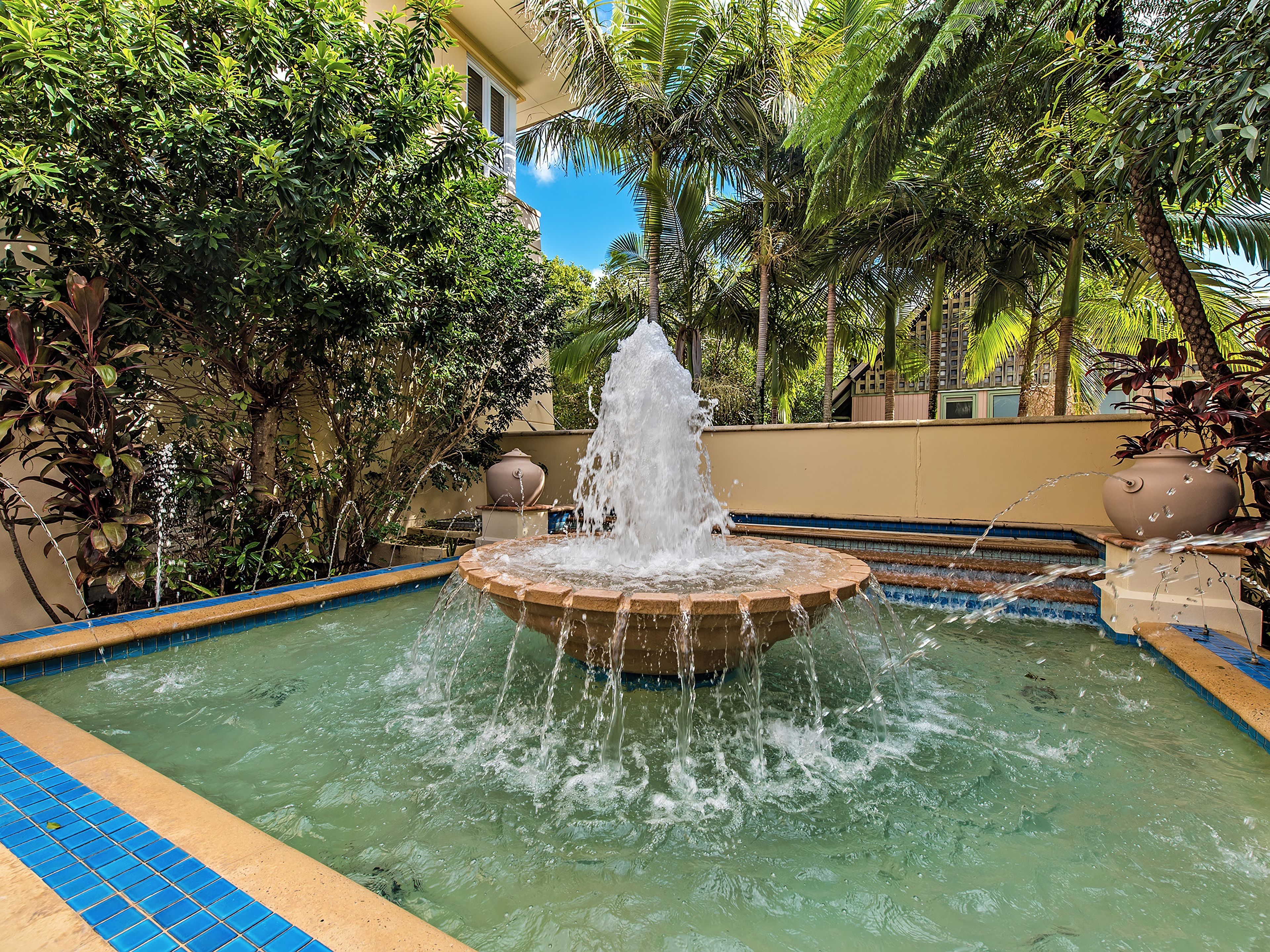 A pool with a central fountain surrounded by lush greenery and a tiled edge.