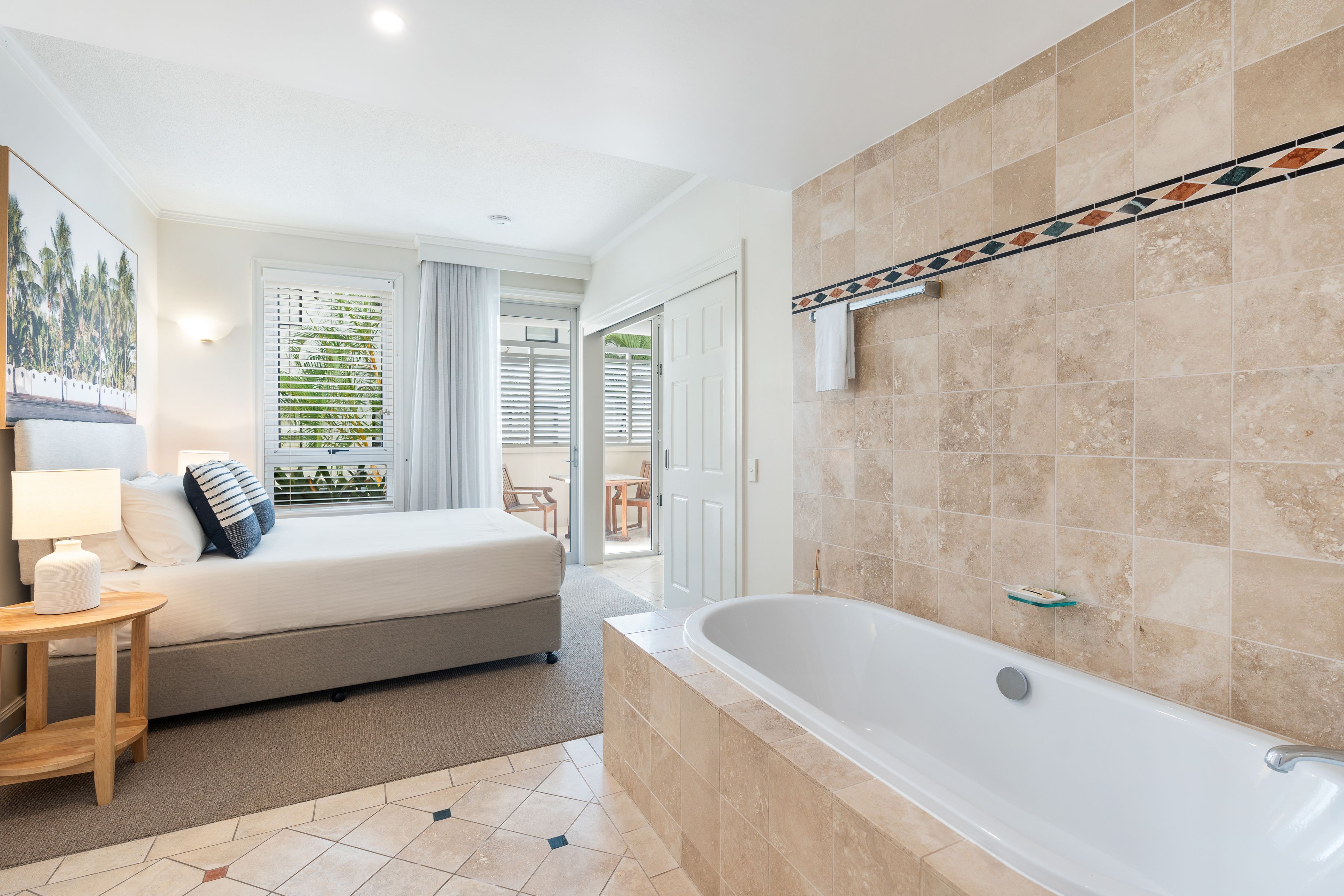 A hotel room with a bed, a bedside table with a lamp, a bathroom with a bathtub, and a view of the outside.
