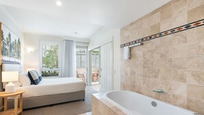 Habitación de hotel con cama, mesita de noche con lámpara, baño con bañera y vistas al exterior.