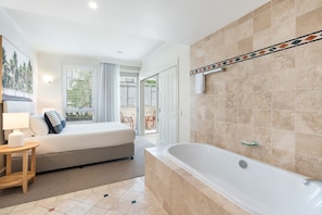 Habitación de hotel con cama, mesita de noche con lámpara, baño con bañera y vistas al exterior.