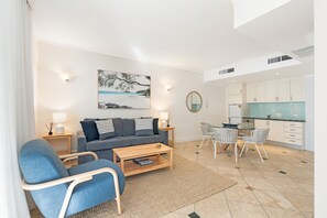 Uma sala de estar moderna com sofá, mesa de centro e cadeiras, uma área de jantar com mesa de vidro e uma cozinha com armários brancos.