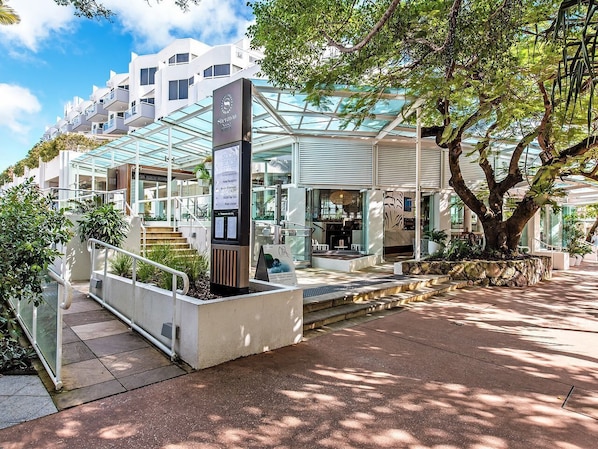 Un hôtel moderne doté d’une façade en verre, d’un espace extérieur aménagé et d’un jardinière en pierre où est planté un arbre.