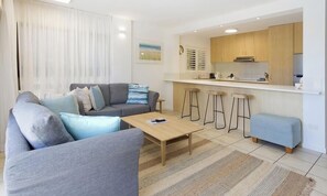 A modern living room with a grey sofa, a wooden coffee table, and a small blue ottoman. A kitchen with wooden cabinets and a bar stool is visible in the background. - Sundown Lodge 1 Edgar Bennett Avenue 14 (Noosa Heads)