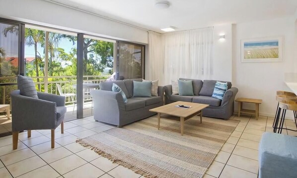 A modern living room with grey sofas, a wooden coffee table, and large windows with a view of greenery.