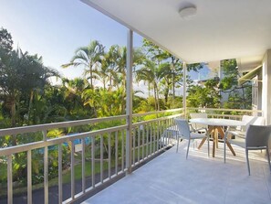 Un balcone con tavolo e sedie, circondato da un parapetto, affacciato su un rigoglioso giardino con palme.