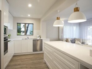 Una cucina moderna con mobili bianchi, un'acciaio inossidabile lavastoviglie e una grande isola con lavello. Sopra l'isola ci sono lampade a sospensione e una finestra con vista sul verde circostante.