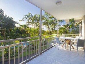 Un balcone con ringhiera bianca, un tavolo e sedie, affacciato su un rigoglioso giardino con diverse piante e alberi.