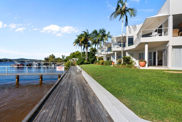 A waterfront property with a wooden dock, a grassy lawn, and a building with balconies overlooking the water.