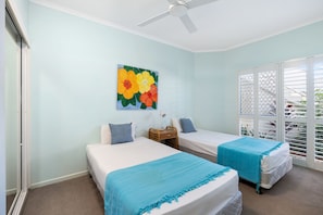 A room with two beds, a ceiling fan, a painting of flowers, and a window with blinds.