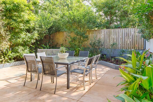An outdoor patio with a glass-top table and chairs, surrounded by lush greenery and a wooden fence.