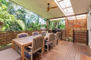 Un espace extérieur abrité, aménagé pour les repas, avec une table et des chaises en bois, un ventilateur de plafond et une vue sur une végétation luxuriante.