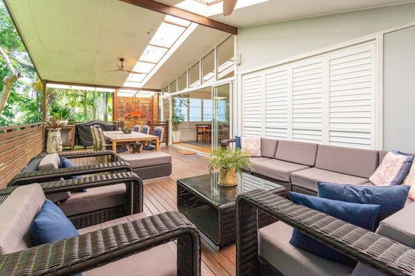 A covered outdoor seating area with wicker furniture, a glass coffee table, and a view of the outdoors.
