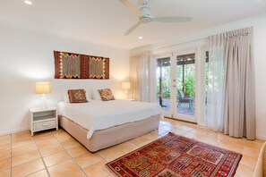 A bedroom with a large bed, two bedside lamps, a ceiling fan, a decorative wall hanging, and a view through a glass door to an outdoor area.