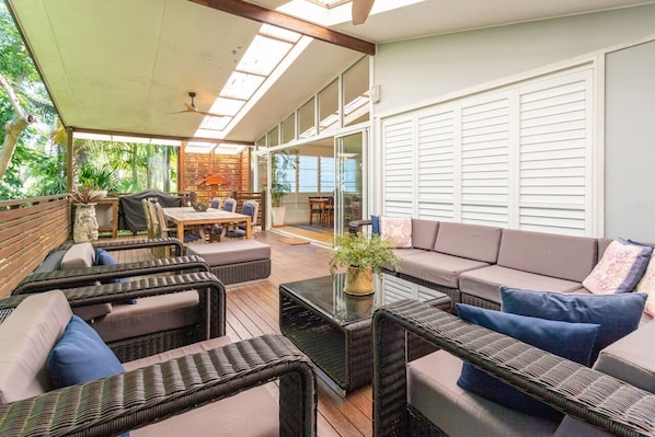 A covered outdoor seating area with wicker furniture, a glass coffee table, and a view of the outdoors.