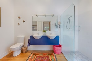 Une salle de bain avec des toilettes blanches, deux lavabos, un miroir, une douche et un tapis coloré.