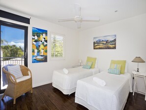 A room with two beds, a wicker chair, a ceiling fan, and artwork on the walls. - Weyba Quays Townhouse 7 Peza Court 6 (Noosa Heads)