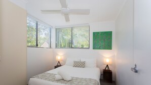 Un dormitorio con una cama, dos mesitas de noche con lámparas, un ventilador de techo y una obra de arte verde en la pared.