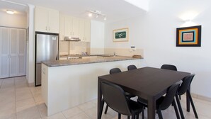 Cocina moderna con armarios blancos, refrigerador de acero inoxidable, horno empotrado y una pequeña isla con pileta. Hay una mesa de comedor con sillas y un cuadro en la pared.