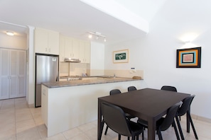 A modern kitchen with white cabinets, a stainless steel refrigerator, a built-in oven, and a small island with a sink. There is a dining table with chairs and a framed picture on the wall. - Headland Views 5 Alderly Terrace 42 (Noosa Heads)