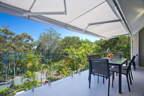 A balcony with a table and chairs, overlooking a lush green landscape with trees and a clear blue sky. - Headland Views 5 Alderly Terrace 42 (Noosa Heads)