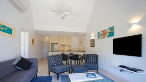 Una sala de estar con sofá, sillas, una mesa de centro y un televisor. Se observa una cocina al fondo.