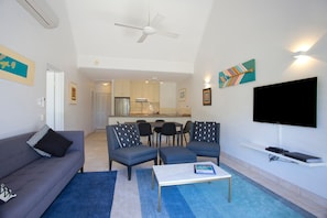 A living room with a sofa, chairs, a coffee table, and a TV. There is a kitchen in the background. - Headland Views 5 Alderly Terrace 42 (Noosa Heads)