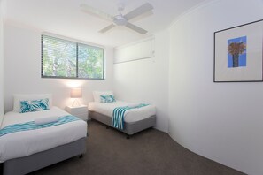 Een slaapkamer met twee bedden, een plafondventilator, een raam met zonwering en een ingelijst kunstwerk aan de muur.