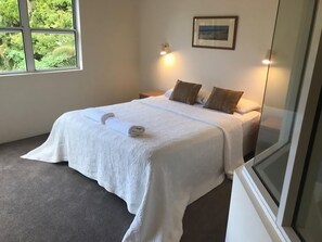 Une chambre à coucher avec un lit, des oreillers, une table de chevet, une lampe fixée au mur et une fenêtre donnant sur un paysage verdoyant.