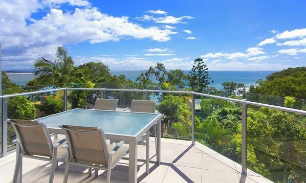 Een balkon met een glazen tafel en stoelen, met uitzicht op een kustlandschap met bomen en de zee.