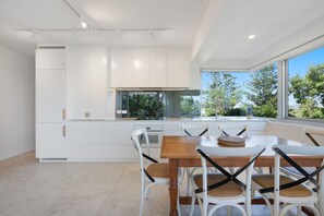 Een moderne keuken met witte kasten, een eettafel met stoelen en uitzicht op groen.