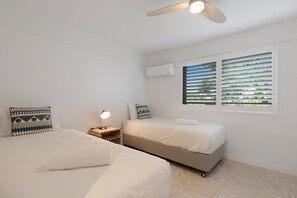 Een slaapkamer met twee bedden, een plafondventilator, een airconditioning en een raam met zonwering.