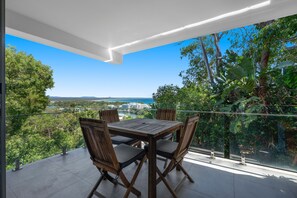 Een balkon met een houten tafel en stoelen, met uitzicht op een kustlandschap met gebouwen en groen.