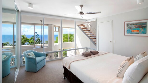 Une chambre moderne avec un grand lit, un fauteuil bleu, un escalier et une vue sur l’océan.