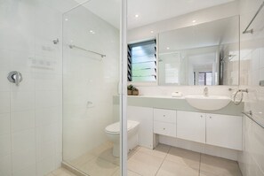 Een moderne badkamer met een glazen douchecabine, een wit toilet, een wastafel met spiegel erboven en een klein raam met jaloezieën.