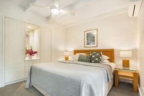 Een hotelkamer met een bed, bedlampjes, een plafondventilator, een nachtkastje, een spiegel en een ingelijste foto aan de muur.