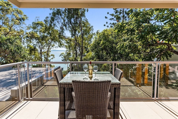 A balcony with a glass table and wicker chairs, overlooking a body of water and trees.