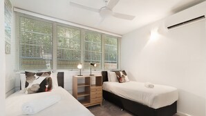 Une chambre avec deux lits, un ventilateur de plafond, une fenêtre avec des stores et une lampe de chevet.