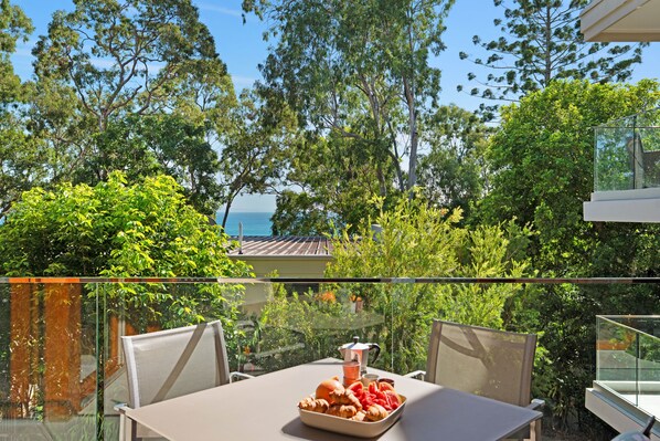 A balcony with a table set for breakfast, overlooking a lush garden and a view of the sea.