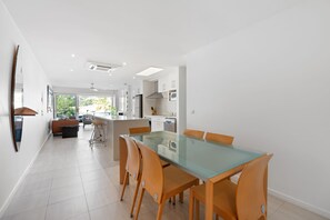 A modern kitchen with white cabinets and stainless steel appliances, a dining area with a glass table and wooden chairs, and a view of the outdoors through large windows. - Jacaranda 12 Hastings Street 12 (Noosa Heads)