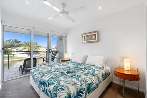 A bedroom with a bed, a chair, a table with a lamp, a ceiling fan, and a view of a body of water. - Jacaranda 12 Hastings Street 12 (Noosa Heads)