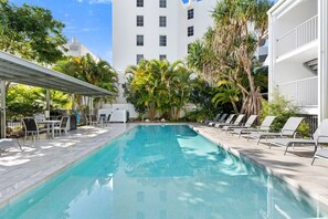A swimming pool with lounge chairs and a covered seating area, surrounded by palm trees and a white building. - Jacaranda 12 Hastings Street 12 (Noosa Heads)