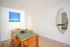 Een eetgedeelte met een glazen tafel, houten stoelen, een schaal met fruit en een schilderij van een strand.