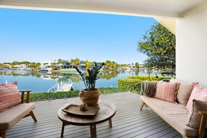 Terraza de madera con un sofá y una mesa, con vistas a un lago tranquilo con barcos, rodeado de vegetación y edificios.