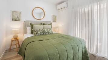 A bedroom with a bed, a round mirror, an air conditioning unit, and a window with sheer curtains.