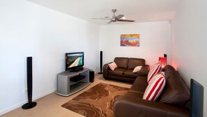 A modern living room with a flat-screen TV, a ceiling fan, a brown leather sofa, and a patterned rug.