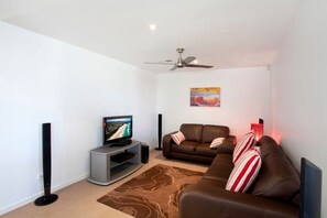 A modern living room with a flat-screen TV, a ceiling fan, a brown leather sofa, and a patterned rug. - Alpha On Elanda - Sunshine Beach Haven (Sunshine Beach)