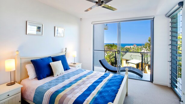 A hotel room with a bed, a ceiling fan, a nightstand with a lamp, a chair, and a balcony with a view of the sea.