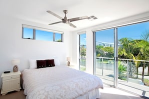 Un dormitorio con cama, mesitas de noche, ventilador de techo y amplias ventanas que dan a vistas de vegetación y un edificio.
