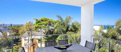Varanda com mesa e cadeiras de madeira, com vista para uma área costeira com árvores e edifícios.