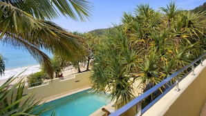 A pool area with a view of the beach, surrounded by palm trees and a clear sky.